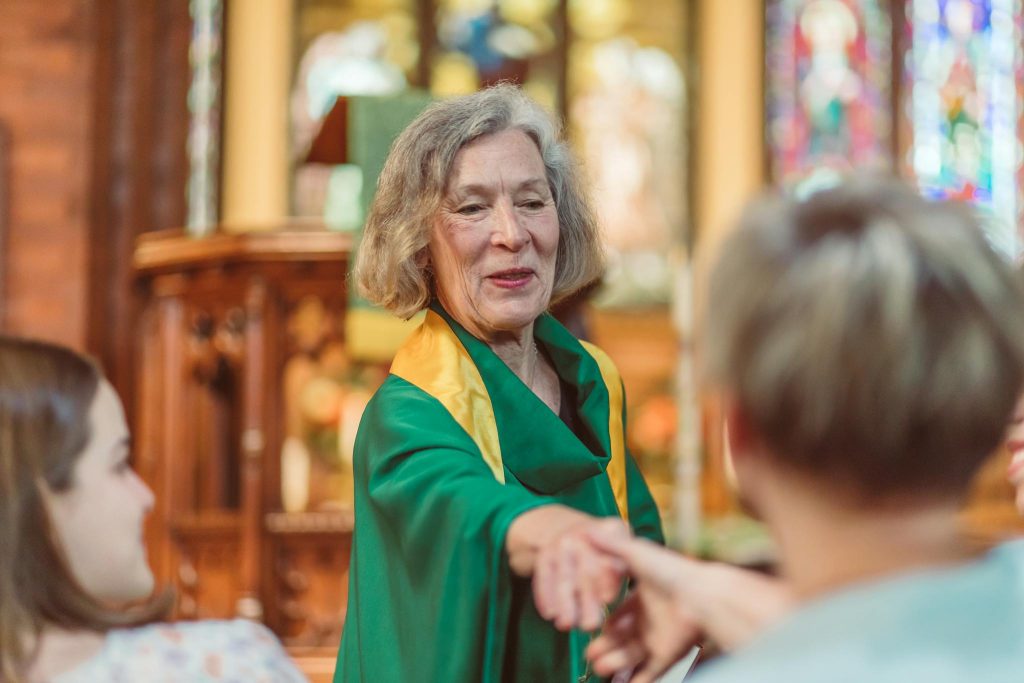 Elderly woman in a green robe engaging with congregants in a vibrant church setting.